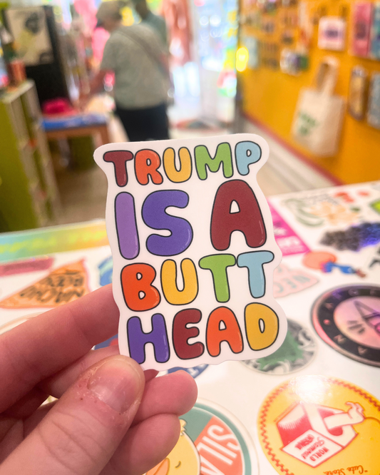Hand holding a colorful vinyl sticker that says “Trump Is A Butthead” in bold rainbow bubble letters. The sticker is 3x2 inches, waterproof, and designed by queer artist Jean-Marie Made It.
