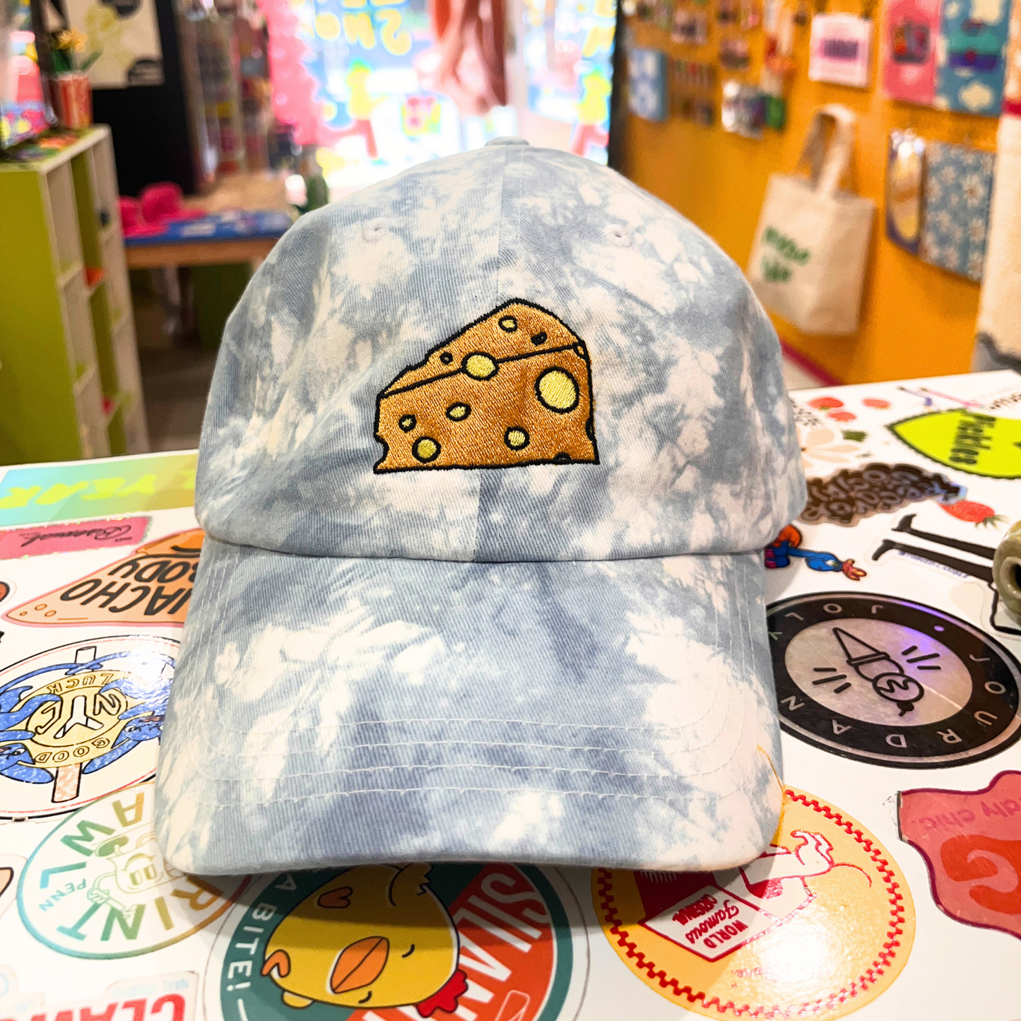 Light blue tie-dye baseball cap with an embroidered cartoon-style wedge of Swiss cheese on the front, displayed inside a colorful, maximalist retail shop.